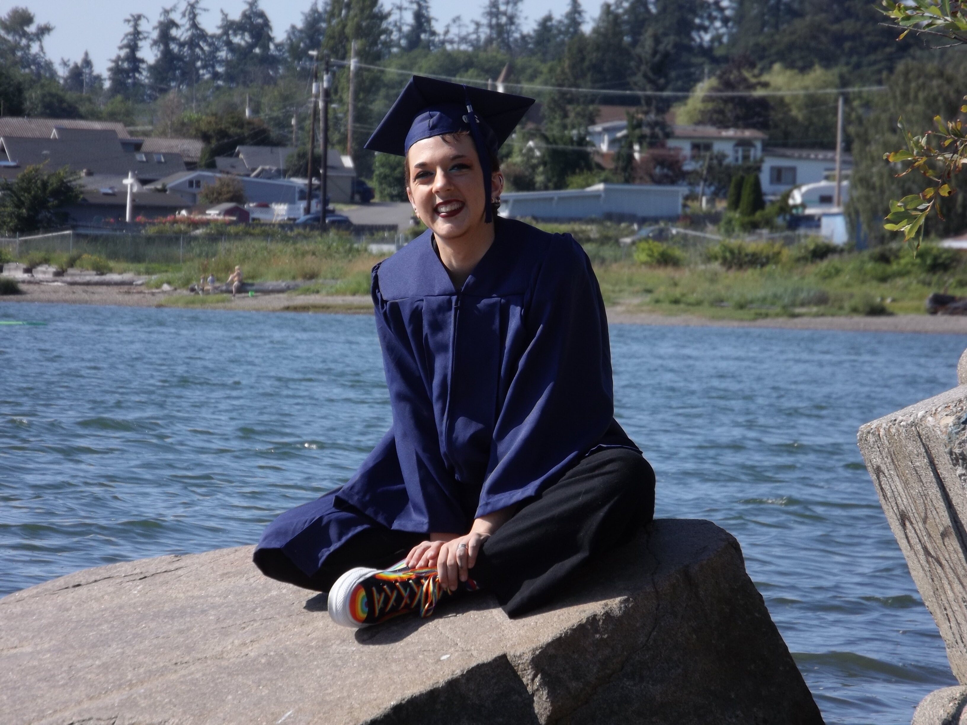 young man sitting rock in front of water, in cap and gown smiling. Student pictured is Tony Void